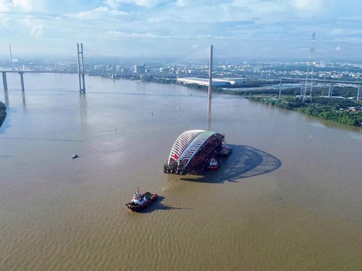 Tàu lai dắt sà lan chở vòm cầu đi bộ trên sông, phía sau là cầu dây văng